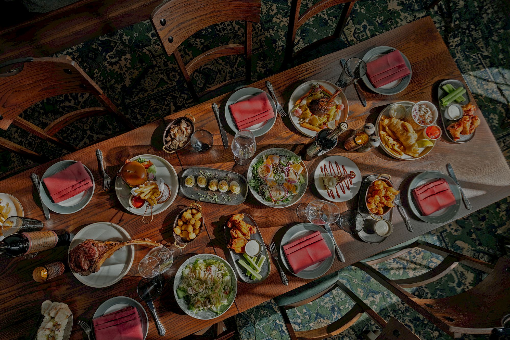 A large dining table is covered with many plated foods, drinks, and red napkins in a cozy restaurant setting, ready for a group meal.