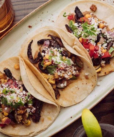 Three tacos topped with veggies and cheese on a plate, plus a lime wedge and drinks nearby, all on a wooden table.