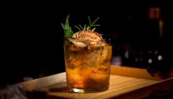 A whiskey or bourbon cocktail on a wooden board, glass filled with ice, garnished with citrus slice and a sprig of herbs, dark bar backdrop.