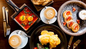 A top-down view of a breakfast spread: shakshuka in a skillet, poached eggs with hollandaise, a pastry, sliced peaches with cream, and coffee cups.