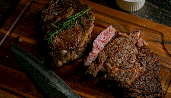 Two medium-rare steaks sliced on a wooden board with a sprig of rosemary and a small ramekin nearby.