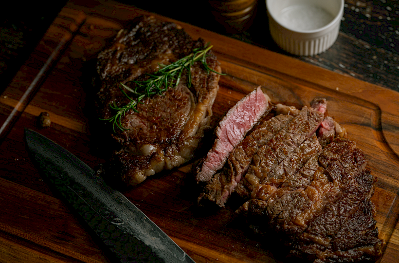 Two medium-rare steaks sliced on a wooden board with a sprig of rosemary and a small ramekin nearby.