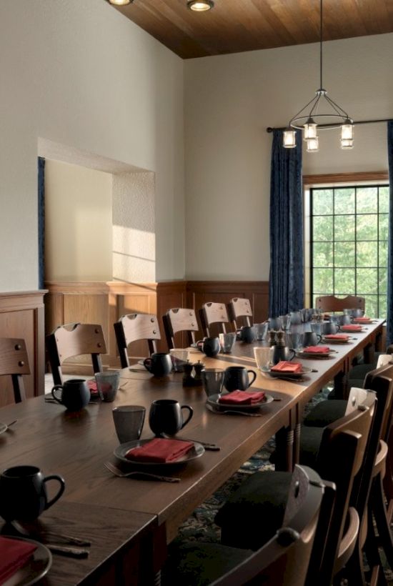 A bright dining room with a long wooden table set for tea, chairs, blue curtains, and a big window.