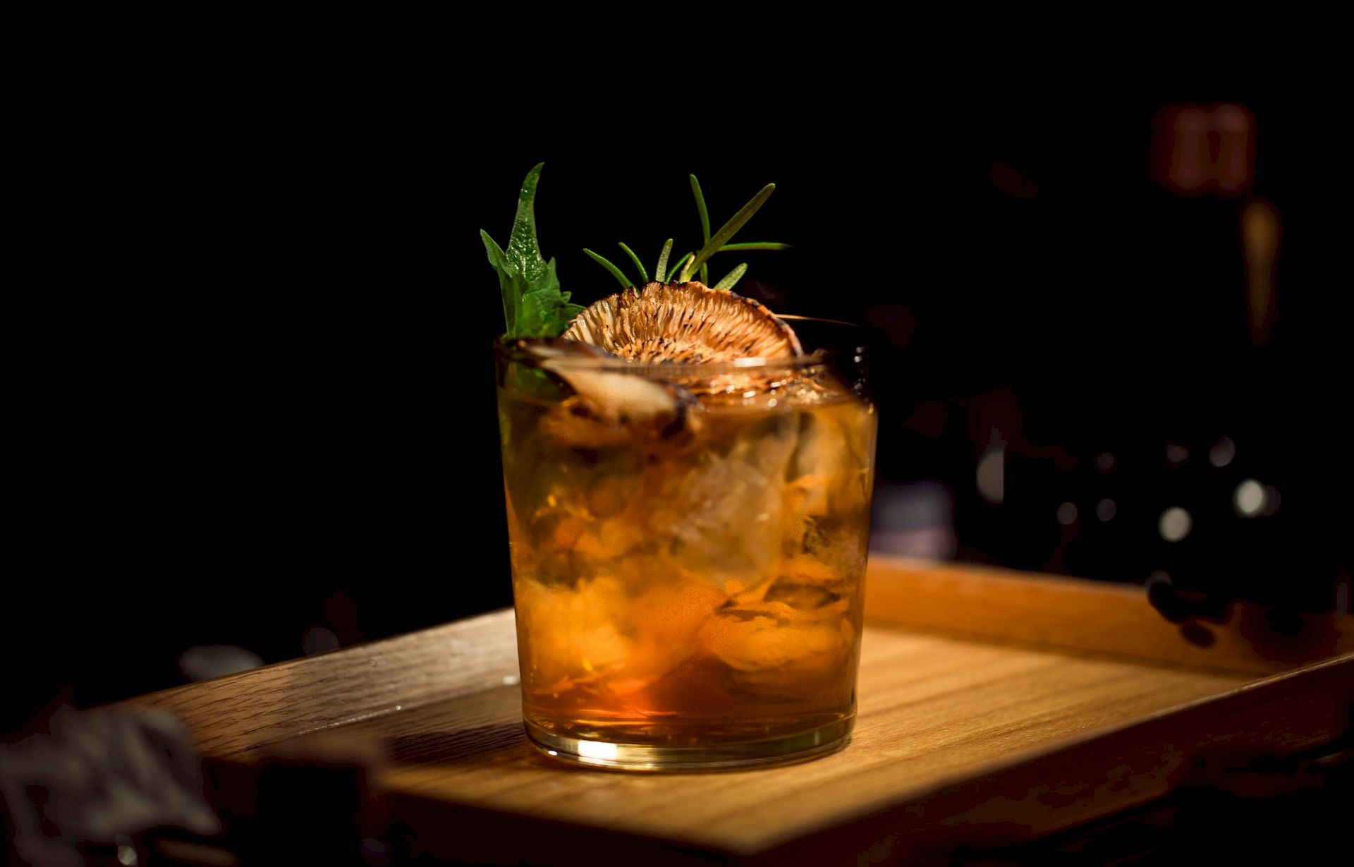 A glass of amber cocktail with ice, garnished with a pineapple slice and a sprig of mint on a wooden board, dimly lit backdrop.