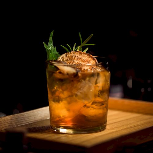 A whiskey cocktail on a wooden board with ice, garnished with herbs and a citrus slice, in a dimly lit bar setting.