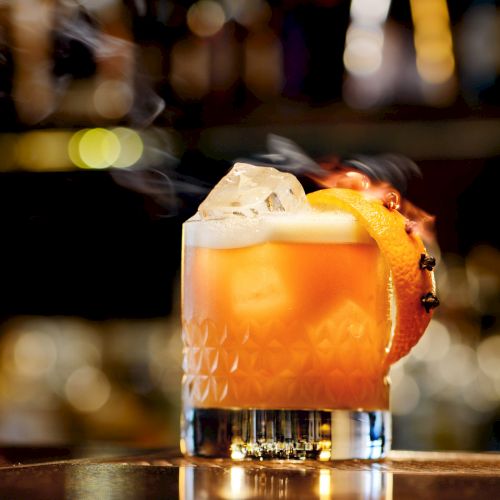 A stylish amber cocktail with ice, a citrus slice garnish, and a frothy top in a short glass at a dimly lit bar.