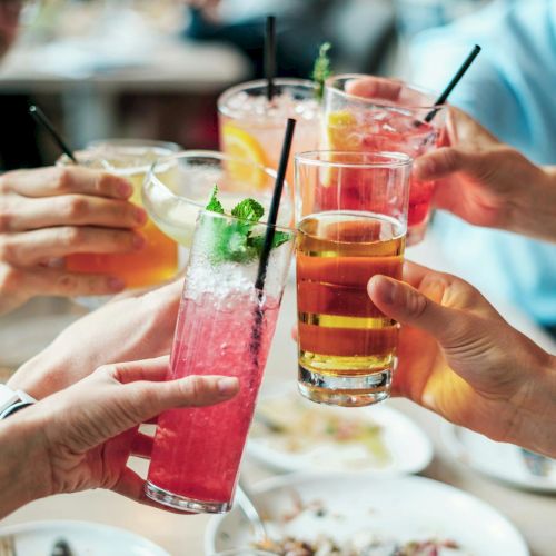 Friends raising colorful drinks in a toast at a crowded table, celebration vibes and clinking glasses. Top it at 140 characters, always ending the sentence.
