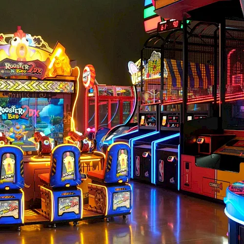 A bright arcade with glowing crane and basketball games, colorful prizes, and neon rides lining the arcade floor.