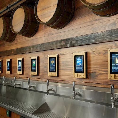 A row of digital taps on a wooden wall with barrels above and green ivy, each screen likely showing beverage options, sleek metal faucet area.