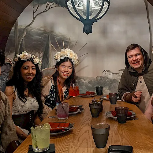 A group of five friends in festive winter outfits and antlers sit at a table with drinks and food, smiling for a photo.